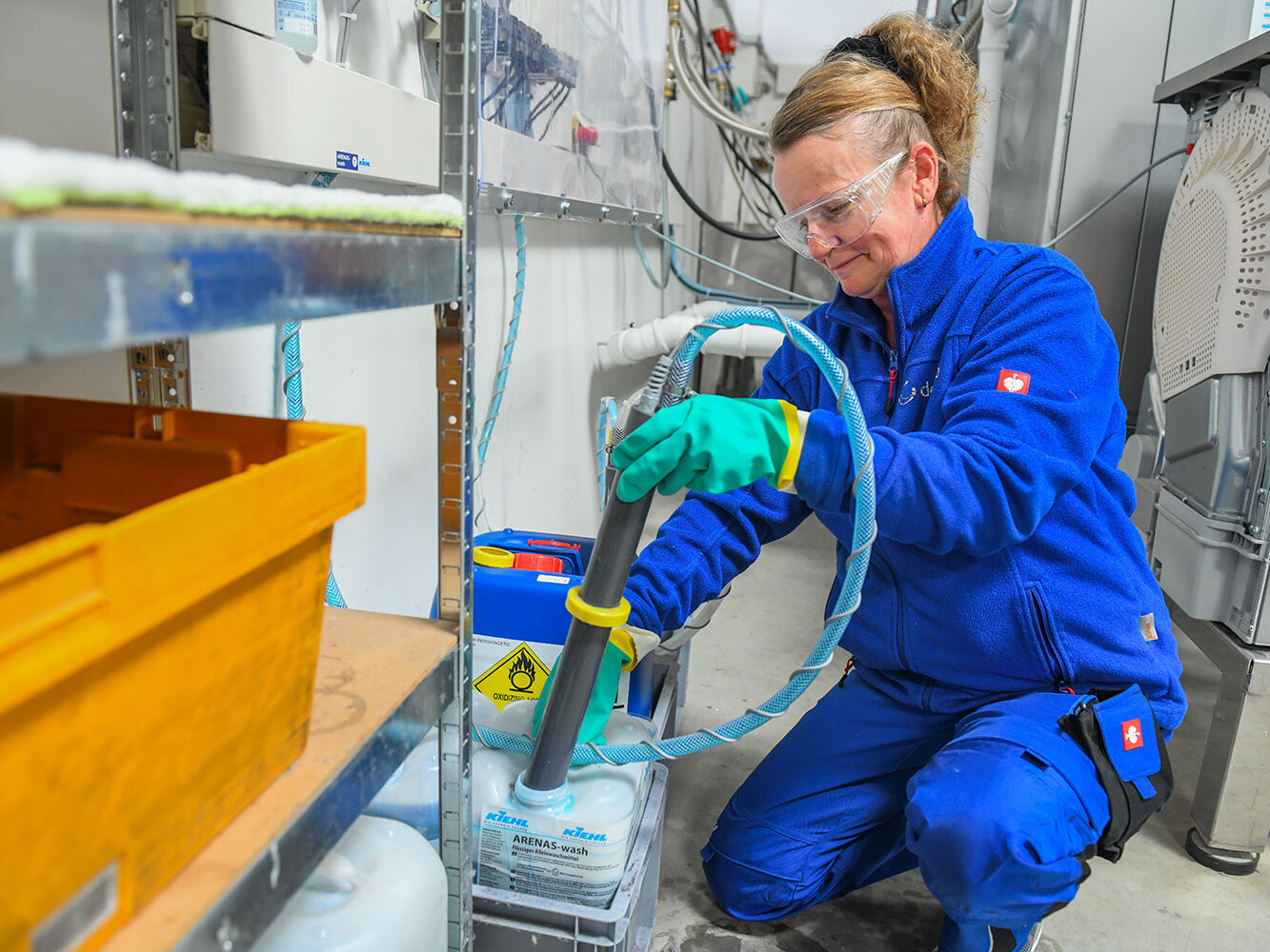 Eine Mitarbeiterin in Schutzbrille und Handschuhen füllt Reinigungsmittel mithilfe eines Schlauches in einen großen Kanister und arbeitet konzentriert im Lagerbereich.