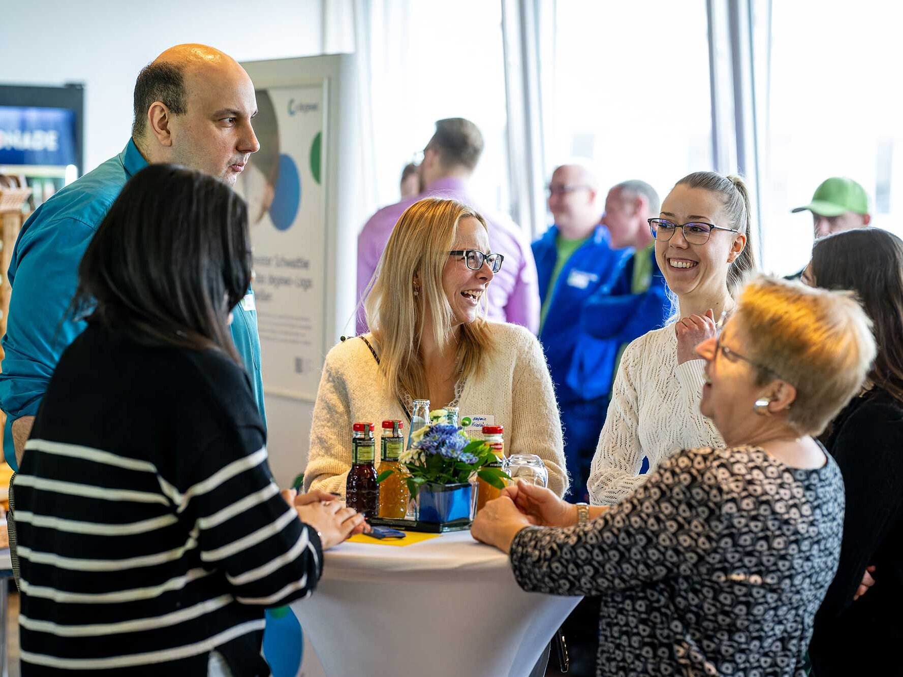 Mehrere Personen stehen an einem runden Stehtisch mit weißer Tischdecke und unterhalten sich. Auf dem Tisch stehen Getränke und eine kleine Blumenvase. Im Hintergrund sind weitere Teilnehmende sowie ein Informationsaufsteller zu sehen. Der Raum ist hell und wirkt wie ein Foyer oder Veranstaltungsbereich.