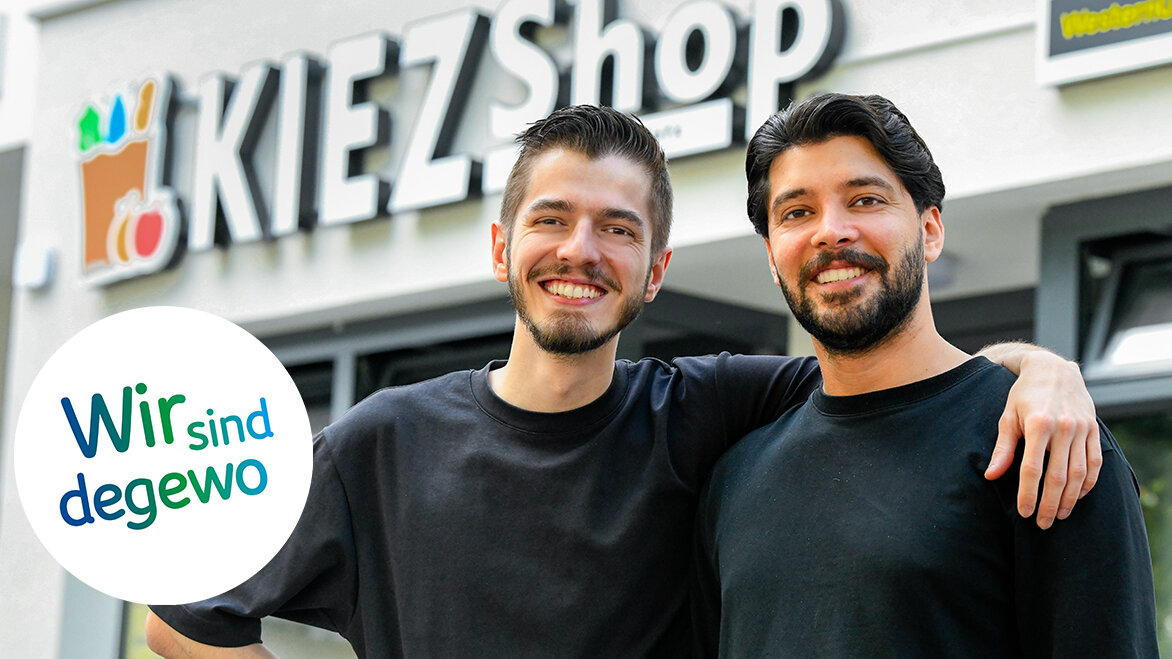 Zwei Männer stehen vor dem Eingang eines Kiezshops im Brunnenviertel in Berlin. Der eine Mann hat seinen Arm um die Schulter des anderen Mannes gelegt. Sie lächeln in die Kamera.