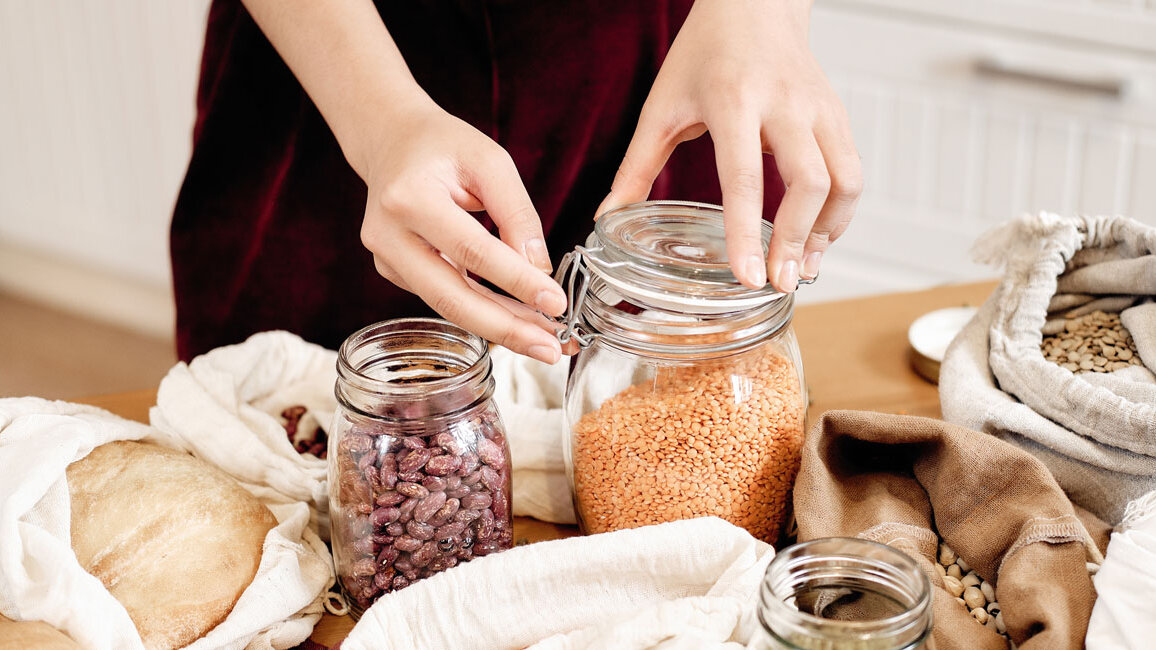 Eine Person schließt ein Einmachglas mit Linsen. Daneben weitere Gläser und Jutesäcke mit Hülsenfrüchten, Nüssen und Brot.