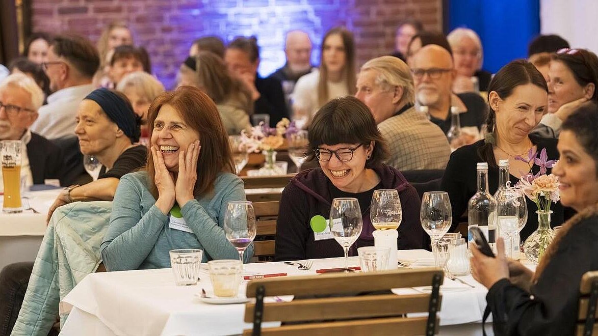 Teilnehmende Mietergremien sitzen am Tisch und lachen herzhaft. Sie schauen dem Bühnenprogramm zu.