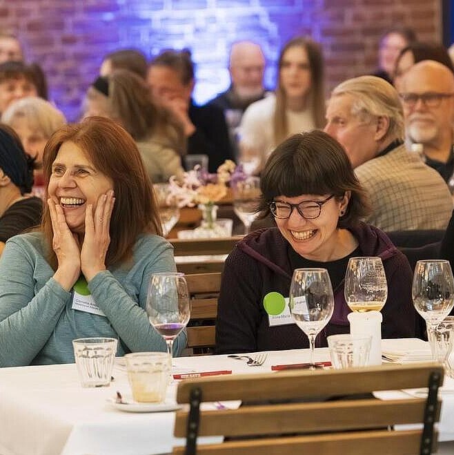 Teilnehmende Mietergremien sitzen am Tisch und lachen herzhaft. Sie schauen dem Bühnenprogramm zu.