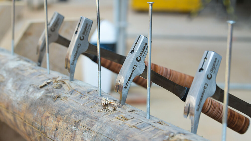 In einem Baumstamm sind fünf Nägel eingeschlagen. Vier Hämmer mit Gravuren von degewo und Schrobsdorff sind ebenso ins Holz geschlagen. Im Hintergrund ist verschwommen eine Baustelle zu sehen.