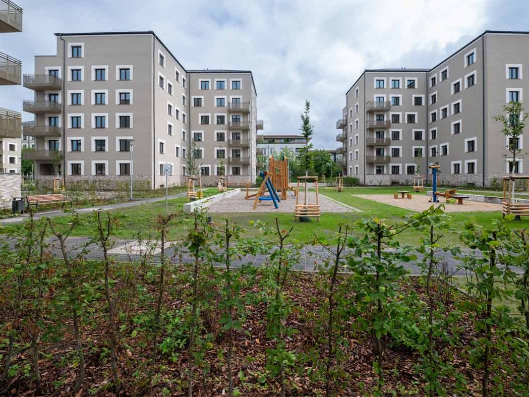 Außenansicht degewo-Neubau Falkenstraße Adlerstraße: Hofseite mit Spielplatz, mit Sträuchern bepflanztes Beet im Vordergrund.