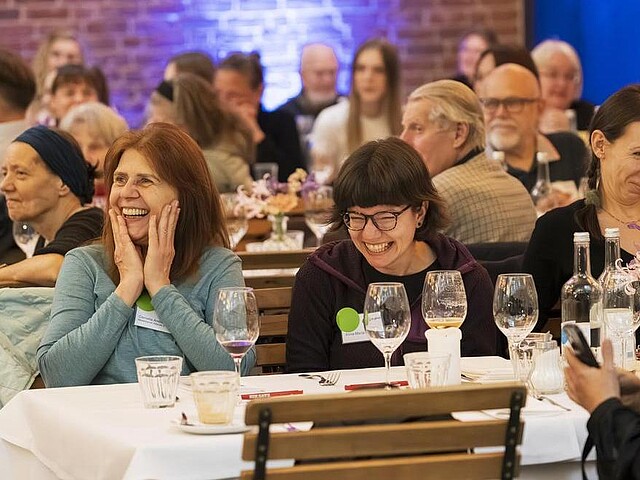 Teilnehmende Mietergremien sitzen am Tisch und lachen herzhaft. Sie schauen dem Bühnenprogramm zu.