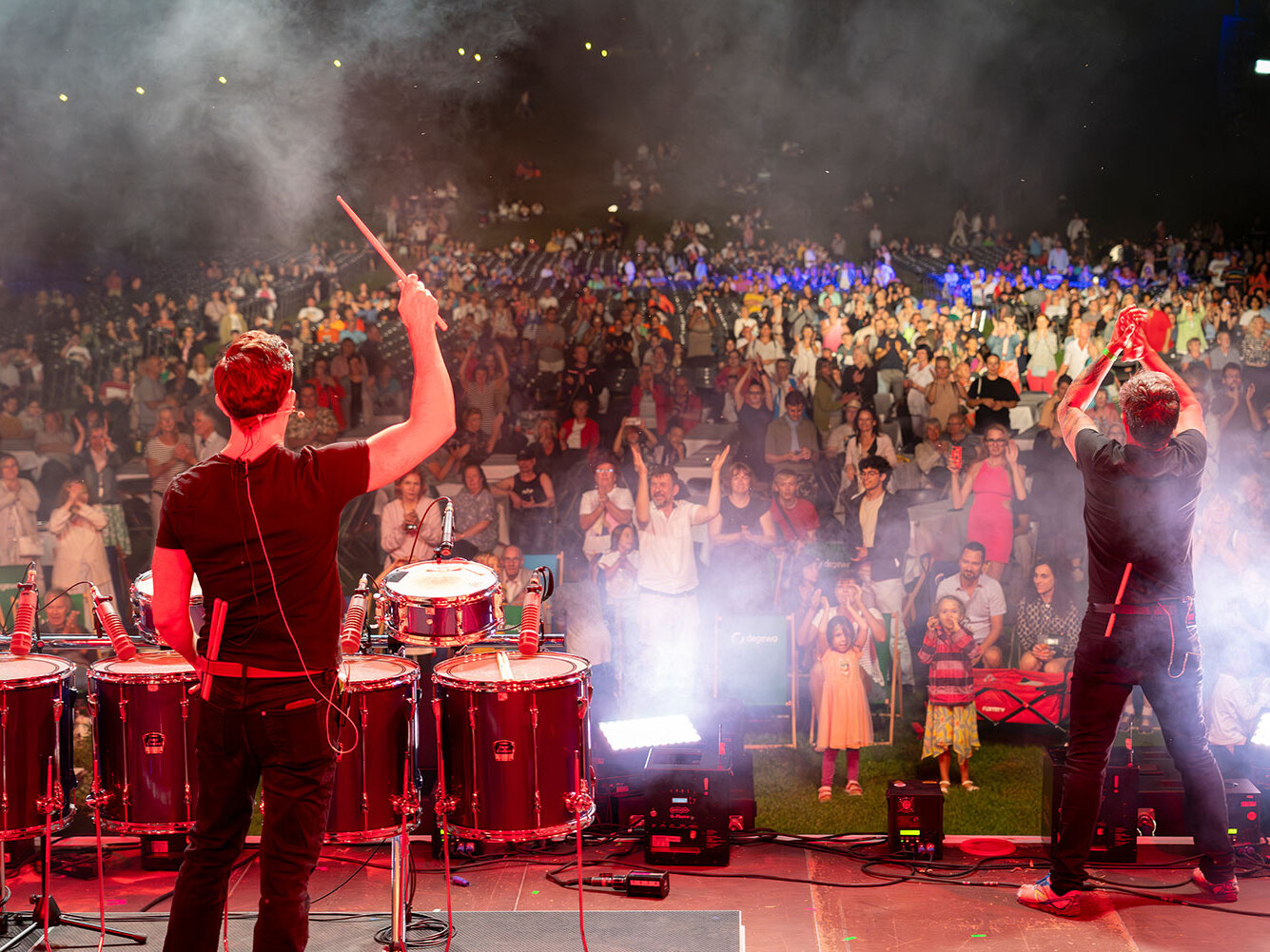 Zwei Musiker stehen auf einer Bühne vor einem großen begeisterten Publikum während Nebel und Lichtstimmung das Konzert untermalen.
