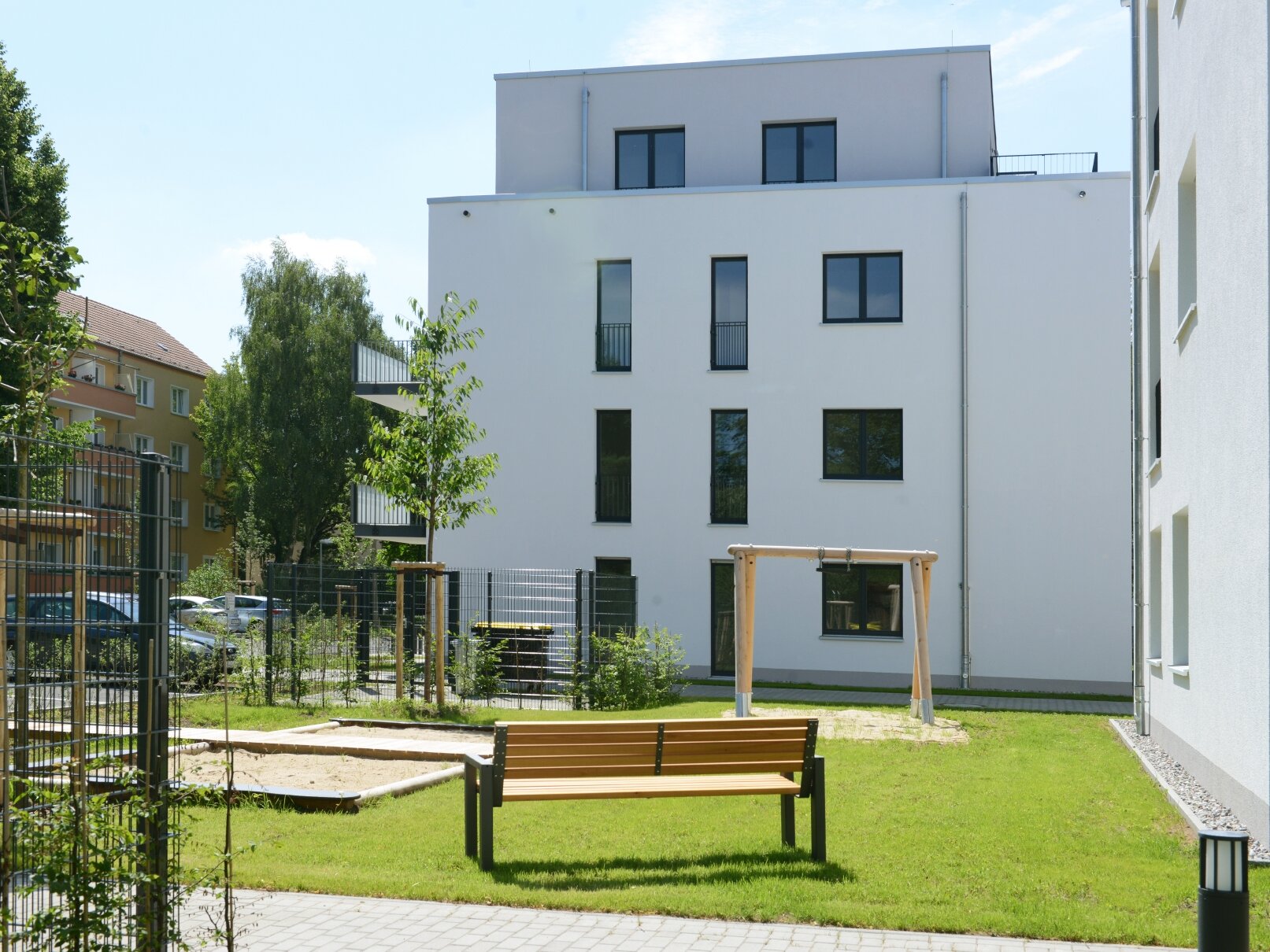 Hofansicht degewo-Neubau Karl-Frank-Straße 33 und 35 mit Zaun, Weg, Weglaterne, Sitzbank und Rasenspielplatz im Vordergrund.