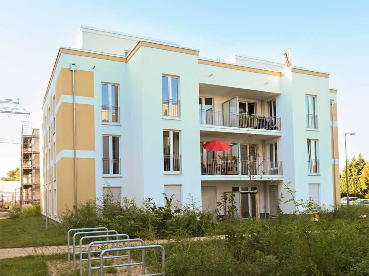 Schrägansicht eines degewo-Neubaus am Eisenhutweg mit drei Vollgeschossen und Staffelgeschoss. Die Fassade ist hell gestaltet und mit gelben Akzentflächen gegliedert. Balkone mit Metallgeländern öffnen sich zur begrünten Außenanlage; im Vordergrund sind Fahrradständer und Rasenflächen zu sehen