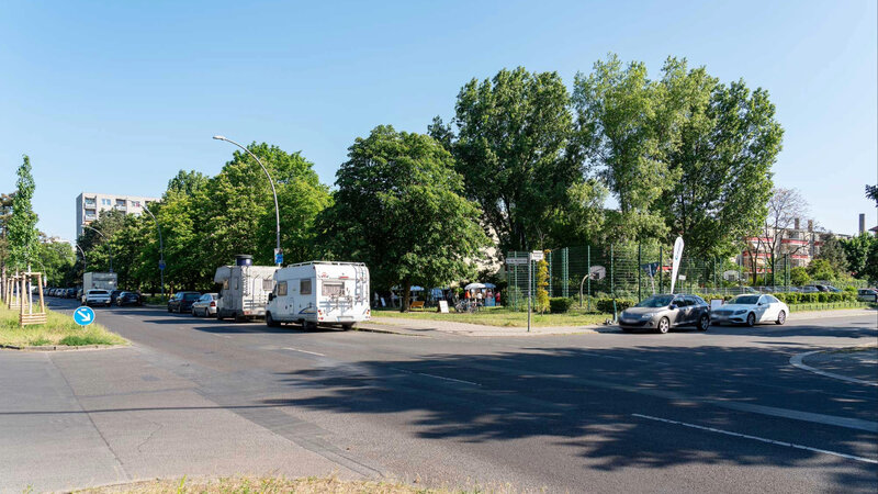 Straßenkreuzung in einem grünen Wohngebiet mit parkenden Autos, Wohnmobilen und Bäumen. Am Straßenrand liegt eine eingezäunte Sport- oder Freizeitfläche, im Hintergrund stehen Wohnhäuser unter blauem Himmel.