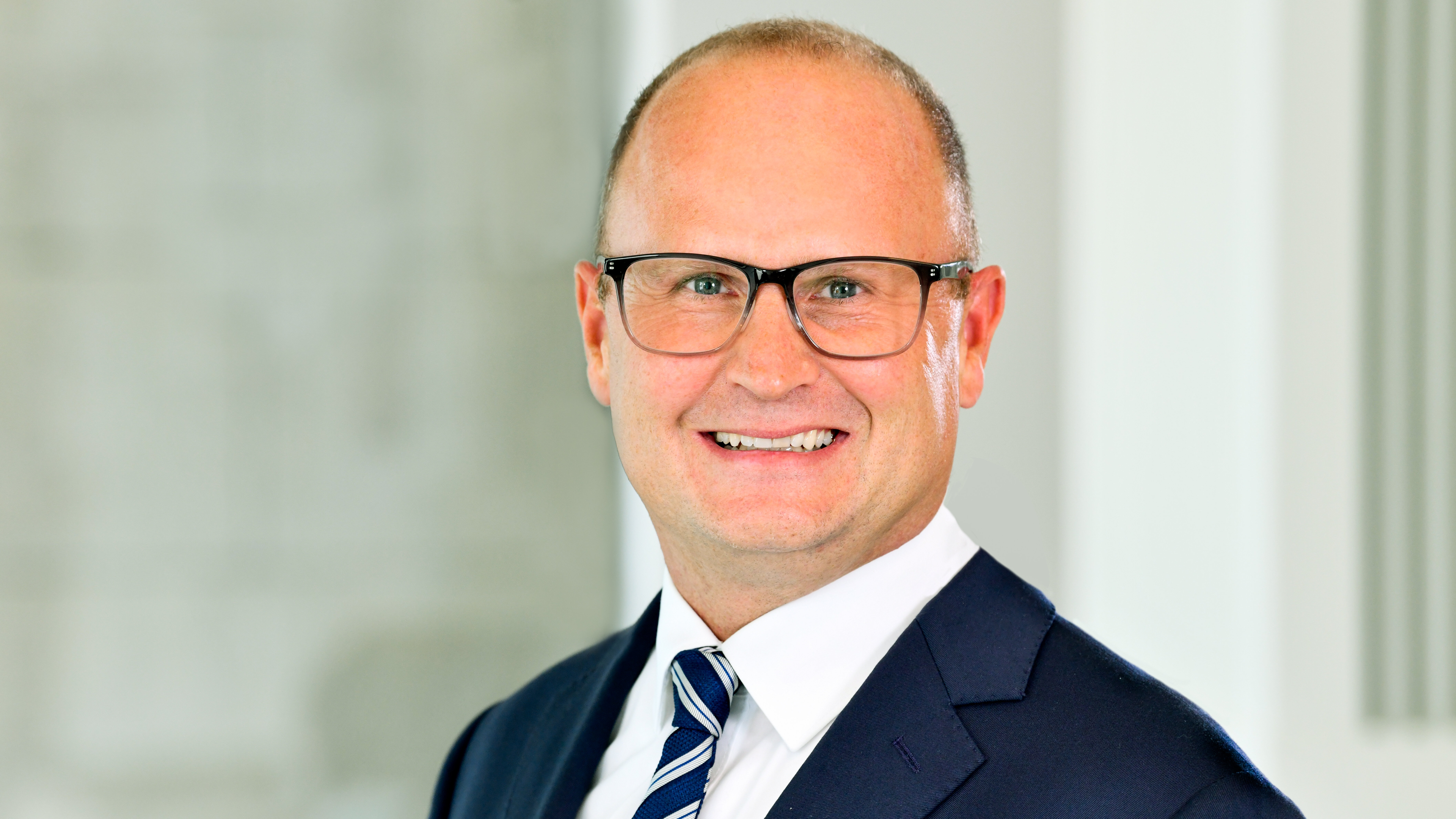 Porträt eines Mannes im dunklen Anzug mit gestreifter Krawatte und Brille vor hellem, weichgezeichnetem Hintergrund. Freundlicher Gesichtsausdruck, frontale Perspektive.