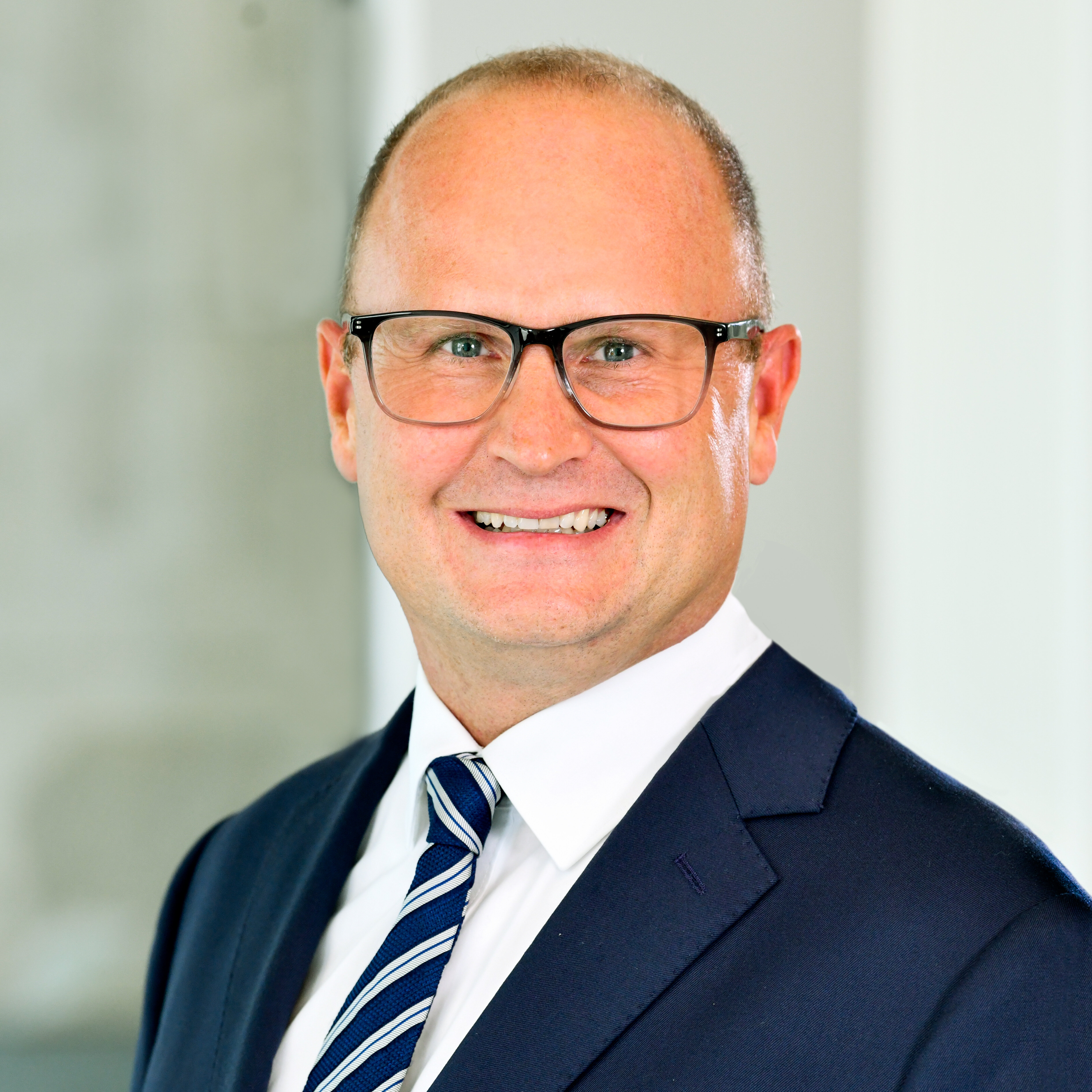 Porträt eines Mannes im dunklen Anzug mit gestreifter Krawatte und Brille vor hellem, weichgezeichnetem Hintergrund. Freundlicher Gesichtsausdruck, frontale Perspektive.