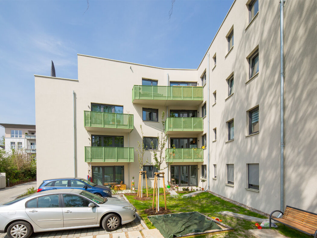 Außenansicht degewo-Neubau Wassersportallee 13 mit Parkflächen, Autos, Grünfläche mit neu gepflanzten Bäumen und Sitzbank im Vordergrund.