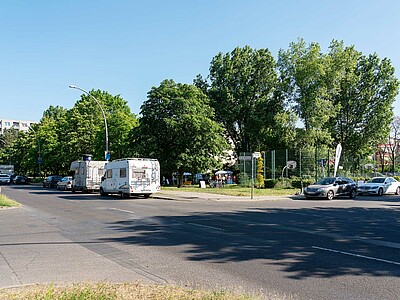 Zu sehen ist eine Straßenkreuzung, am Straßenrand stehen Autos und Wohnmobile. Im Hintergrund sind eine Rasenfläche, Gebäude und ein Sportplatz zu sehen. 