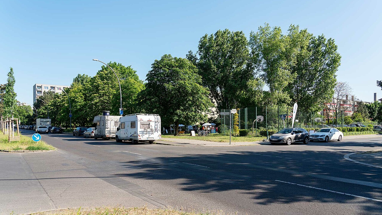Zu sehen ist eine Straßenkreuzung, am Straßenrand stehen Autos und Wohnmobile. Im Hintergrund sind eine Rasenfläche, Gebäude und ein Sportplatz zu sehen. 