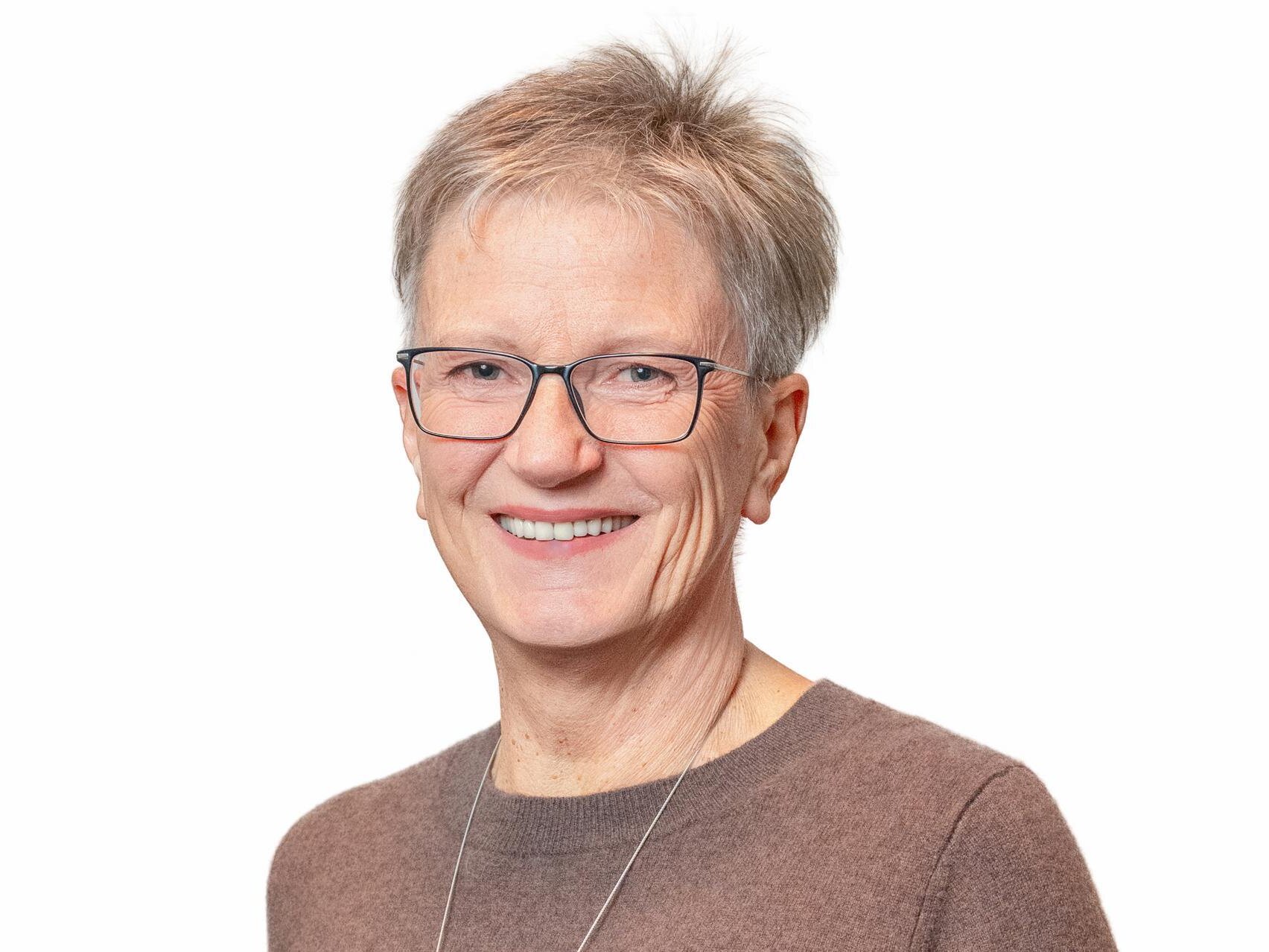 Eine ältere Frau mit kurzem, blondem Haar und einer Brille lächelt freundlich in die Kamera. Sie trägt ein hellbraunes Oberteil.