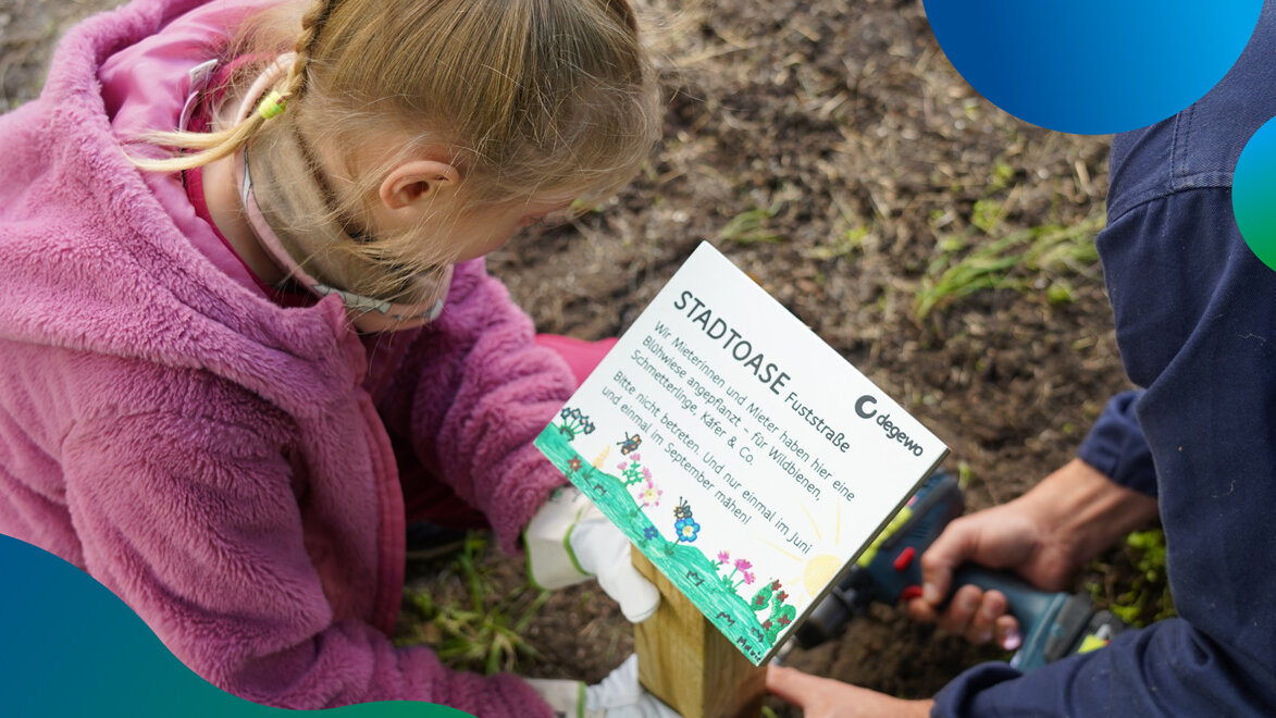 Ein Kind in pinker Jacke befestigt gemeinsam mit einer erwachsenen Person ein Schild im Boden, auf dem "Stadtoase" steht.