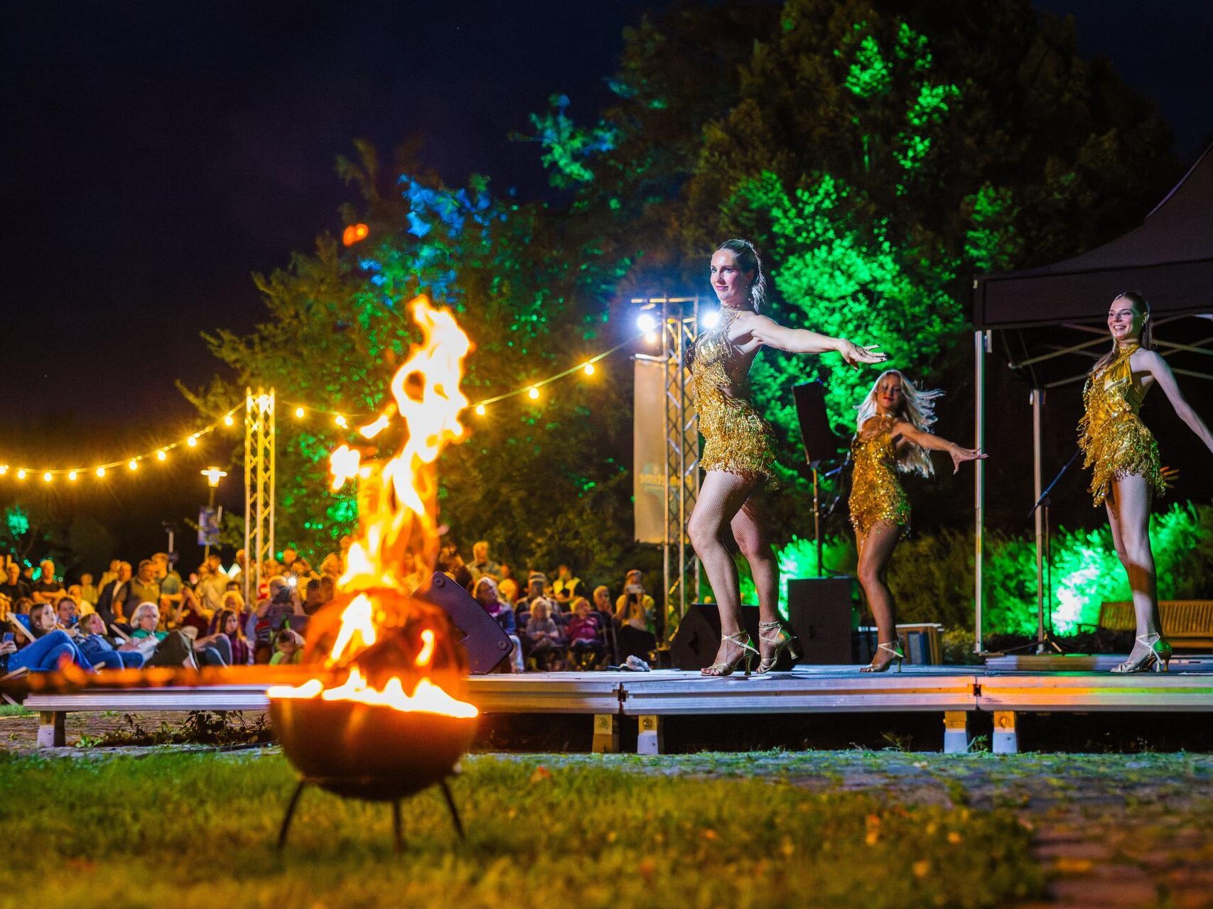 Drei Künstlerinnen und Künstler treten bei Dunkelheit auf einer Open-Air-Bühne auf. Im Vordergrund brennt eine Feuerschale, während das Publikum die Show unter Lichterketten verfolgt.