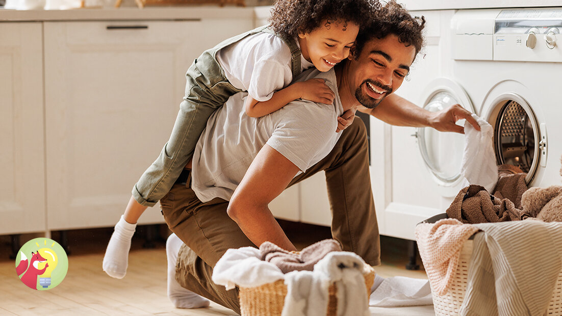 Ein lachender Mann kniet vor einer geöffneten Waschmaschine und hält ein Handtuch in der Hand. Auf seinem Rücken hängt ein lachendes Kind. Vor ihm stehen zwei Wäschekörbe.