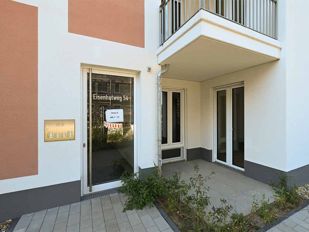Detailansicht des Hauseingangs Eisenhutweg 54 mit verglaster Tür, Klingel- und Briefkastenanlage. Der Eingangsbereich ist barrierearm gestaltet und führt direkt in das moderne Mehrfamilienhaus mit Balkonen und hellen Fassadenflächen.