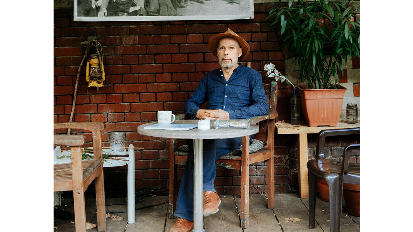 Frontaufnahmen von dem Künstler Thomas Hendriksson. Er sitzt an einem Tisch, darauf liegen verschiedene Sachen. Hinter ihm hängt ein schwarz-weißes Bild