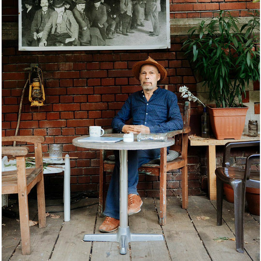 Frontaufnahmen von dem Künstler Thomas Hendriksson. Er sitzt an einem Tisch, darauf liegen verschiedene Sachen. Hinter ihm hängt ein schwarz-weißes Bild