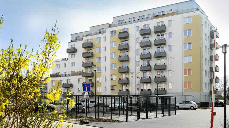 Außenansicht degewo-Neubau Schützenstraße 11 A bis C mit Parkflächen, geöffneter Schranke, Straßenlaterne und blühenden Zweigen links im Vordergrund.