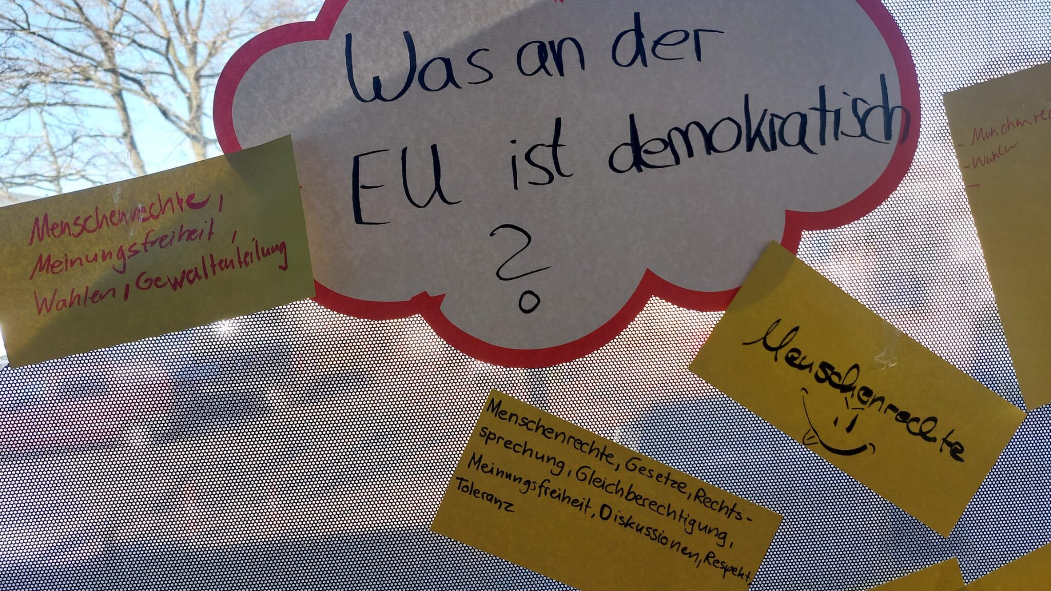 Handgeschriebene Wortwolke am Fenster mit der Frage "Was an der EU ist demokratisch?" und Notizen zu Menschenrechten, Wahlen und Meinungsfreiheit.