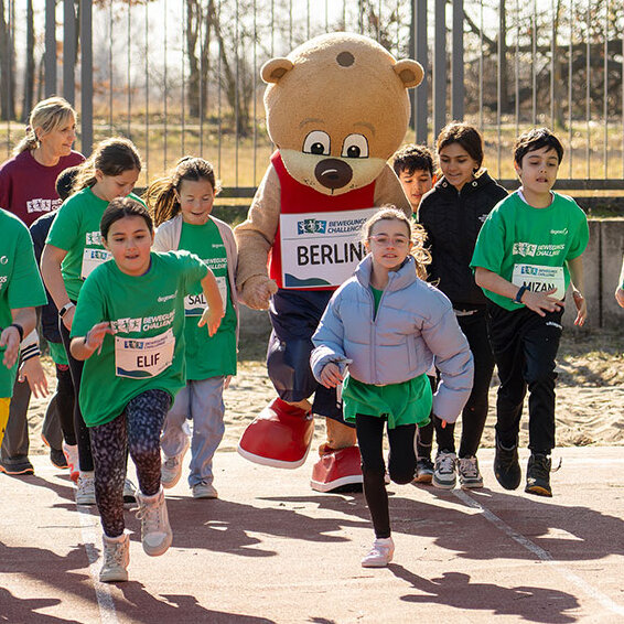 Eine Gruppe Kinder rennt auf einem Sportplatz mit dem Maskottchen Berlino.