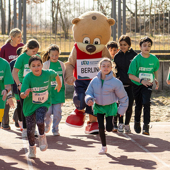 Eine Gruppe Kinder rennt auf einem Sportplatz mit dem Maskottchen Berlino.