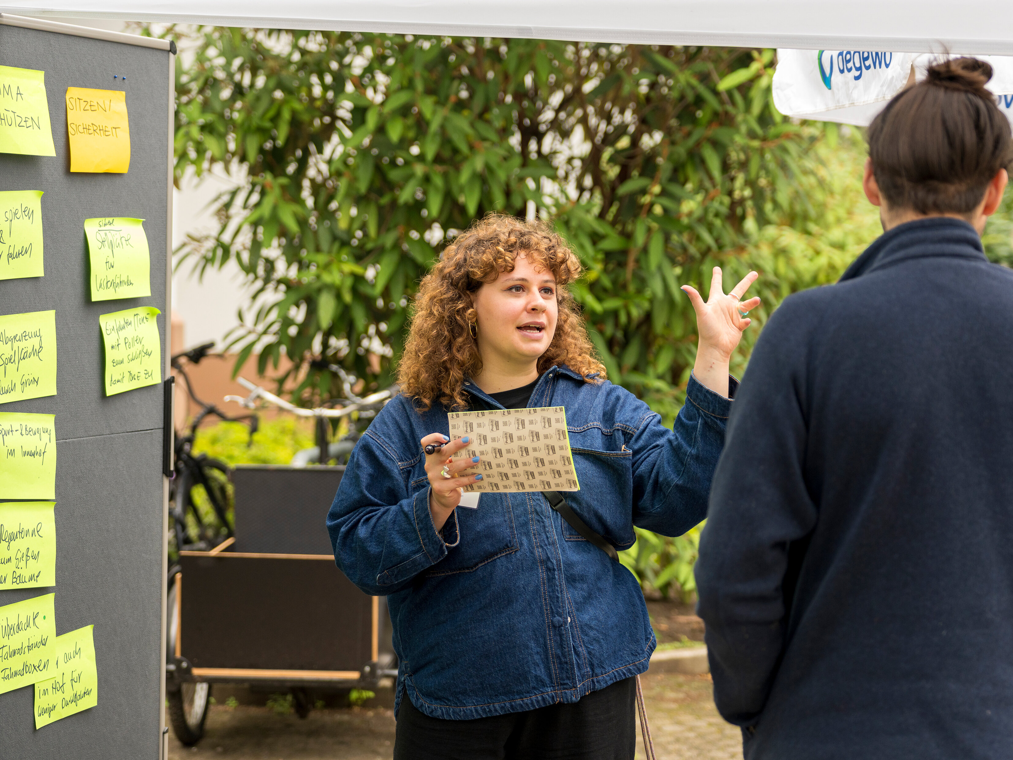 Moderierte Diskussion vor einer Pinnwand mit Haftnotizen zu Themen wie Klima, Sicherheit und Spielflächen. Eine Mitarbeiterin erläutert Inhalte im Gespräch.