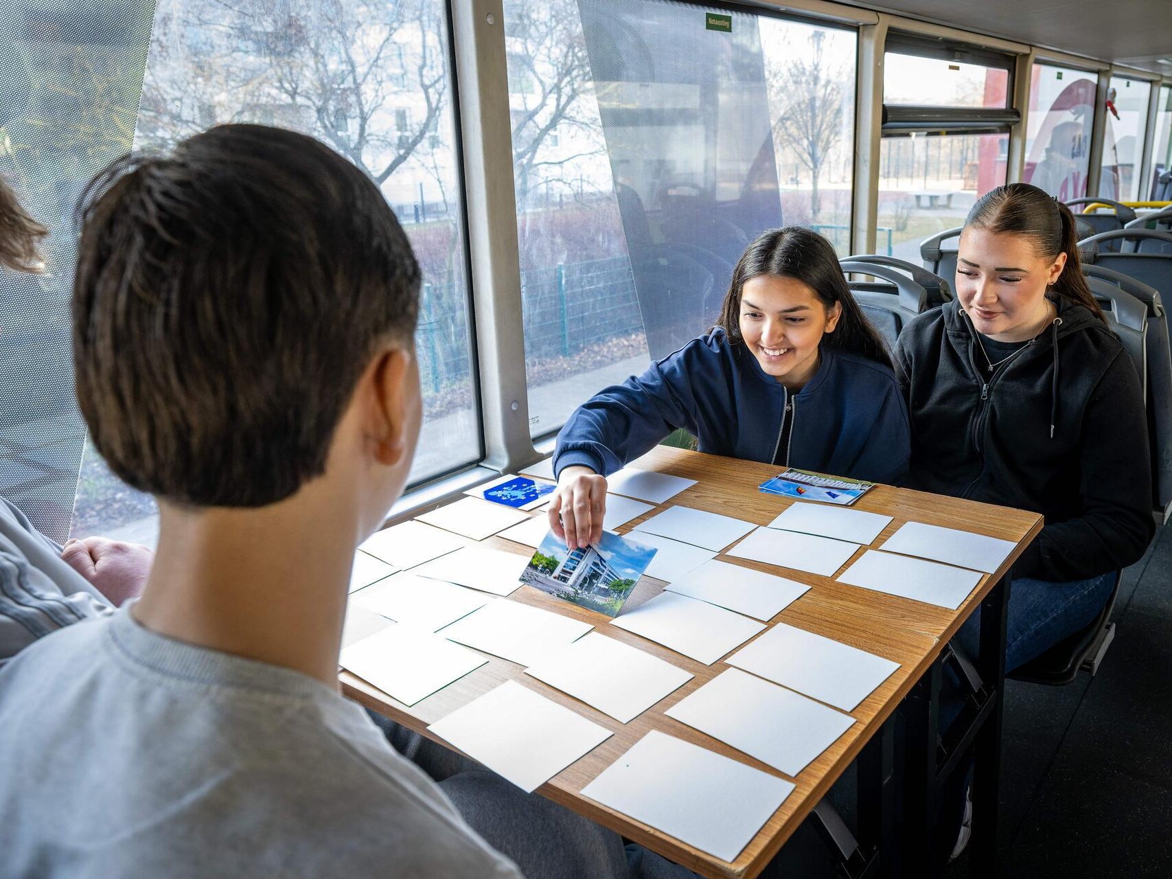 Vier Jugendliche sitze im Demokratie-Mobil um einen Tisch. Auf dem Tisch liegen mehrere umgedrehte Karten. Ein Mädchen dreht eine der Karten um.