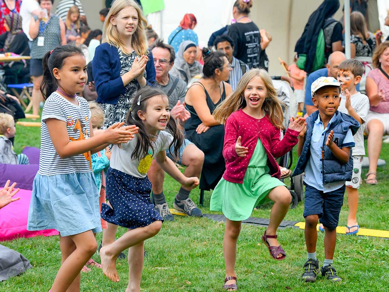 Eine Gruppe fröhlicher Kinder tanzt und spielt auf einer Wiese bei einer Veranstaltung während Erwachsene im Hintergrund zuschauen.