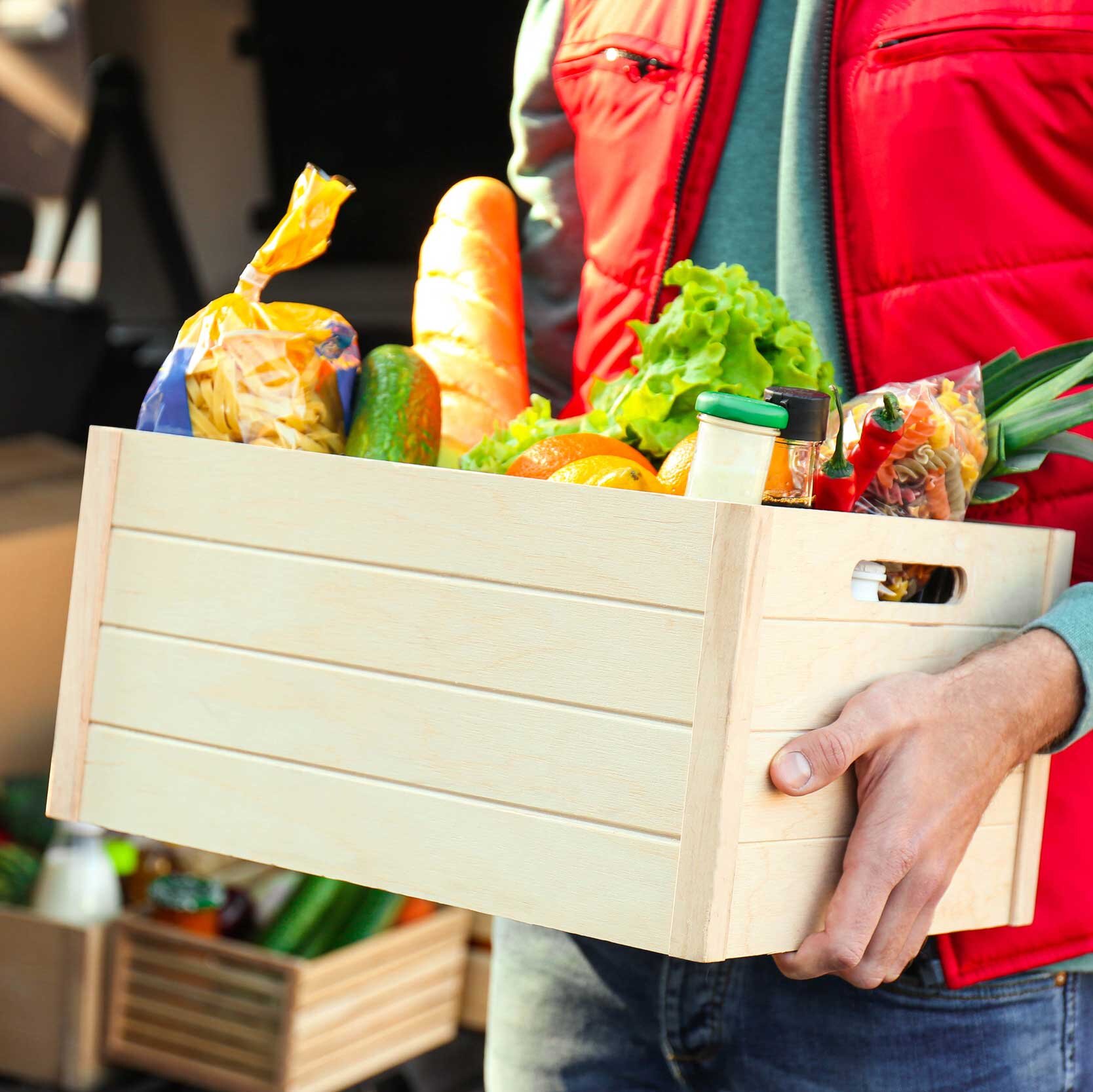 Person in roter Weste trägt eine Holzkiste mit Lebensmitteln wie Brot, Salat, Obst und Gemüse vor einem geöffneten Lieferfahrzeug.