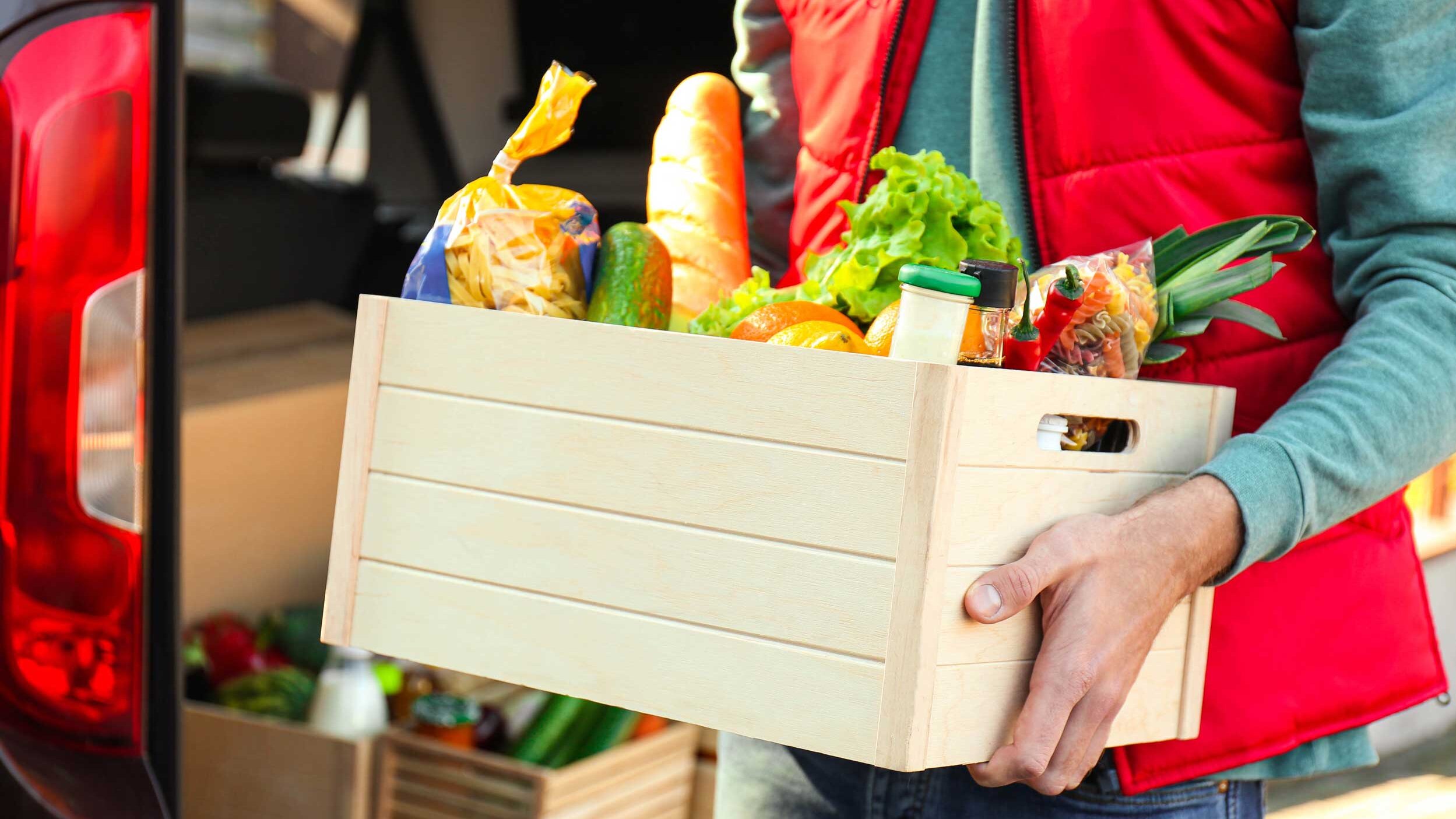 Person in roter Weste trägt eine Holzkiste mit Lebensmitteln wie Brot, Salat, Obst und Gemüse vor einem geöffneten Lieferfahrzeug.