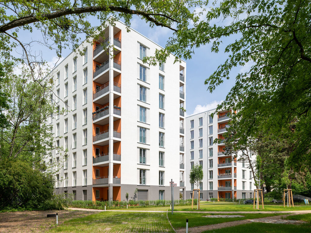 Außenansicht degewo-Neubau Ursulastraße 2 mit Wegen und Rasenflächen sowie links und rechts beblätterten Baumästen und -zweigen im Vordergrund.