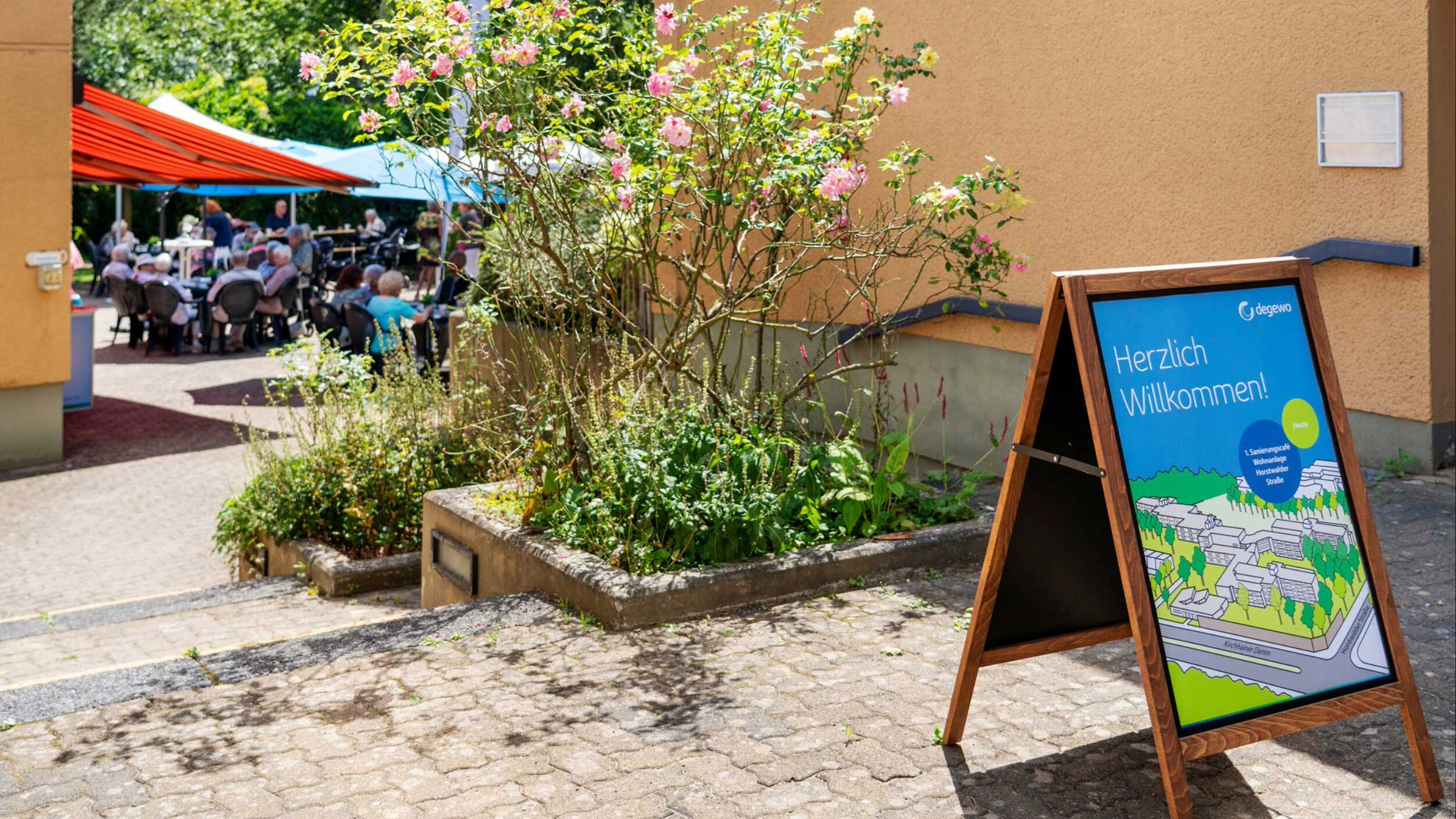 Blick auf das Sanierungscafé in der Wohnanlage Horstwalder Straße: Zahlreiche Bewohnerinnen und Bewohner sitzen an Tischen im Außenbereich, während Projektbeteiligte vor dem Gebäude informieren.