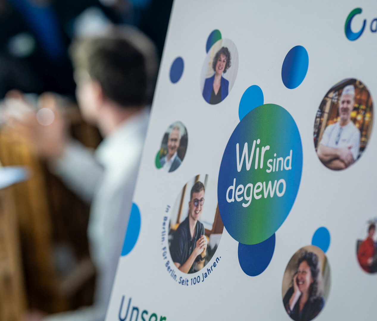 Aufsteller mit Schriftzug Wir sind degewo. Im Hintergrund sind Menschen zu sehen, die ein Foto machen. 