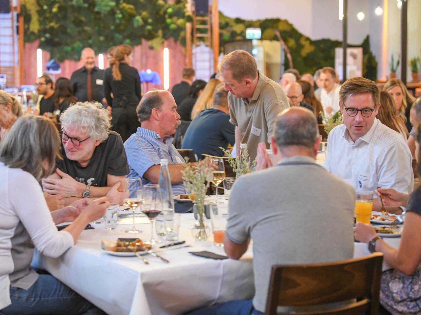 Teilnehmende sitzen an mehreren Tischen in einem Veranstaltungsraum, essen gemeinsam und tauschen sich aus. Im Hintergrund sind weitere Gäste sowie die dekorierte Innenraumgestaltung zu sehen.