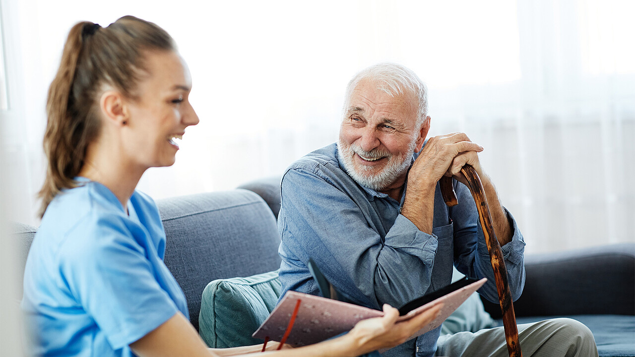 Eine Pflegerin und ein älterer Mann sitzen gemeinsam auf einer Couch und lachen.
