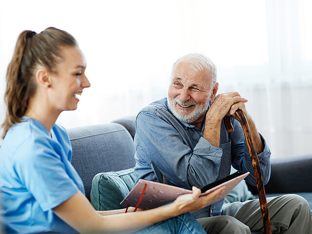 Eine Pflegerin und ein älterer Mann sitzen gemeinsam auf einer Couch und lachen.