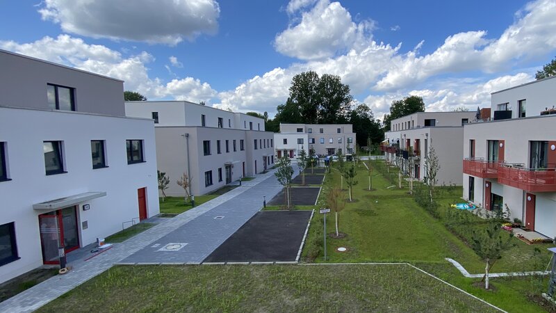 Außenansicht degewo-Neubau Fürstenwalder Allee 324/326: links und rechts Häuser, in der Mitte Grünflächen, neu gepflanzte Bäume sowie links auch Parkflächen und Weg.