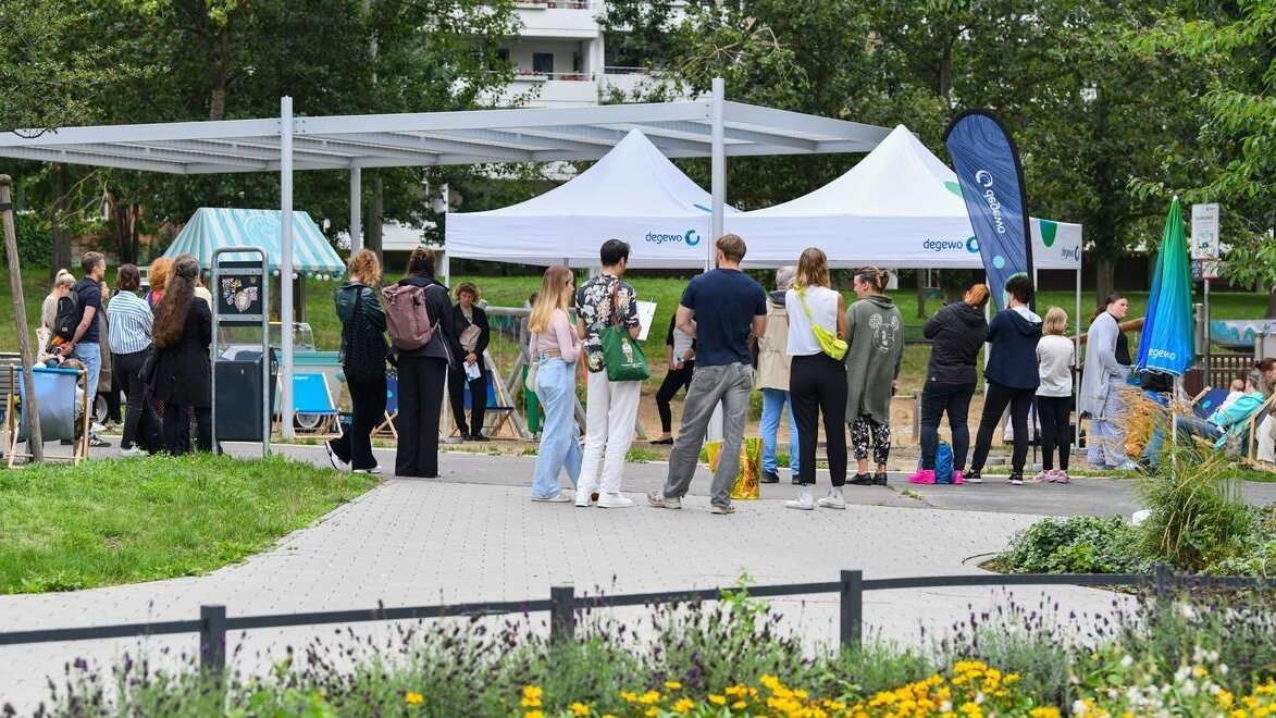 Menschen stehen und gehen auf einem Weg in einer grünen Parkanlage, vor ihnen mehrere weiße Informationszelte mit degewo-Logo; rundherum sind Bäume, Sitzgelegenheiten und bepflanzte Flächen zu sehen.