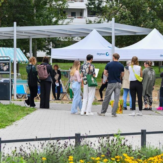 Menschen stehen und gehen auf einem Weg in einer grünen Parkanlage, vor ihnen mehrere weiße Informationszelte mit degewo-Logo; rundherum sind Bäume, Sitzgelegenheiten und bepflanzte Flächen zu sehen.