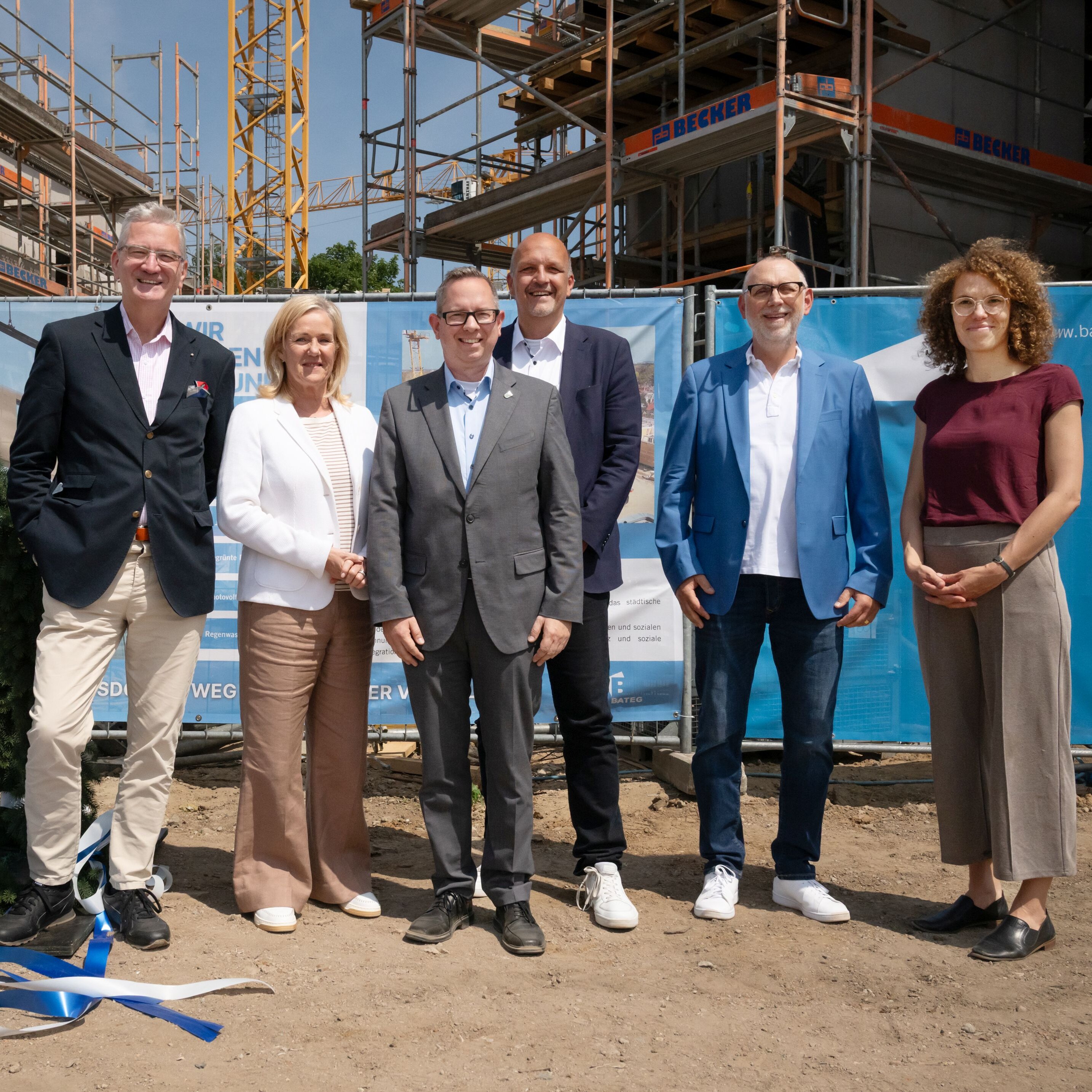 Sechs Personen stehen neben einer Richtkrone vor einer Baustelle mit Bauzaun.