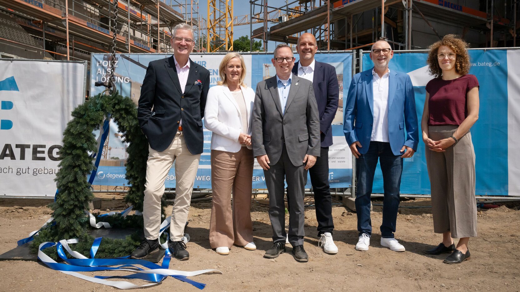 Sechs Personen stehen neben einer Richtkrone vor einer Baustelle mit Bauzaun.