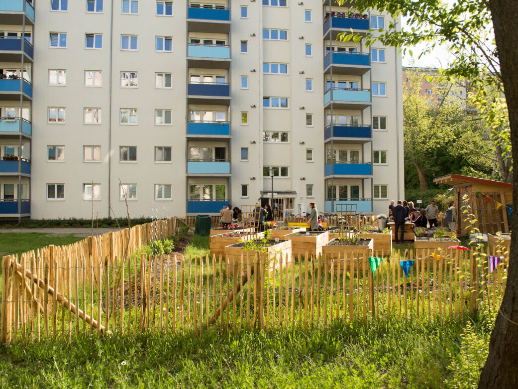 Vor einem mehrstöckigen Wohnhaus ist eine Grünfläche und eingezäunter Garten zu erkennen. In diesem halten sich Mieter des Hauses auf.