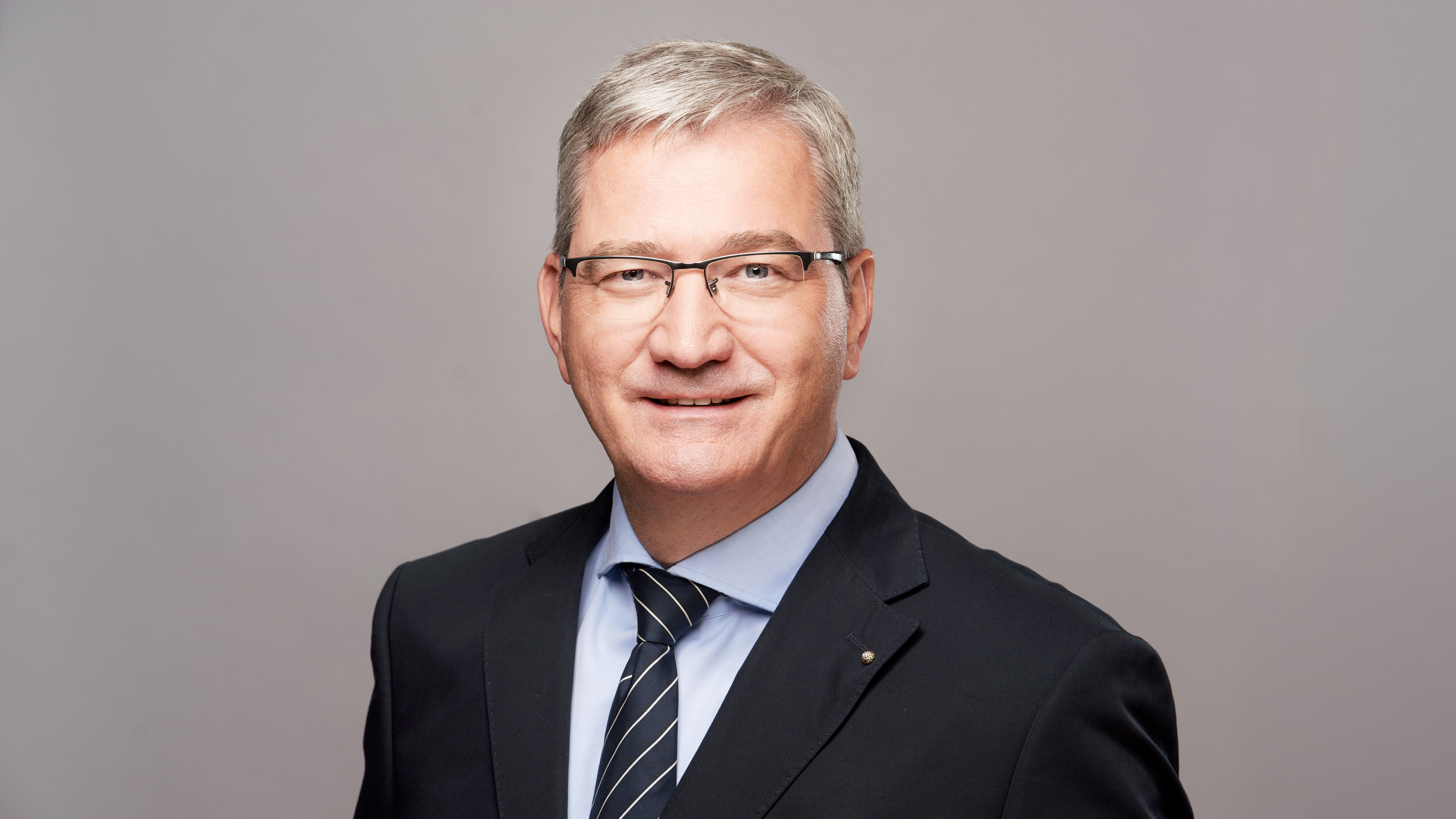 Studioaufnahme eines Mannes im Anzug mit Krawatte und Brille vor neutralem, grauem Hintergrund. Er blickt frontal in die Kamera und lächelt leicht.