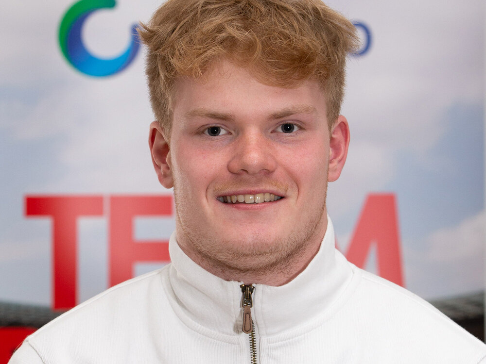 Junger Mann in einem weißen Pullover posiert vor einer Sponsorenwand und schaut direkt in die Kamera.