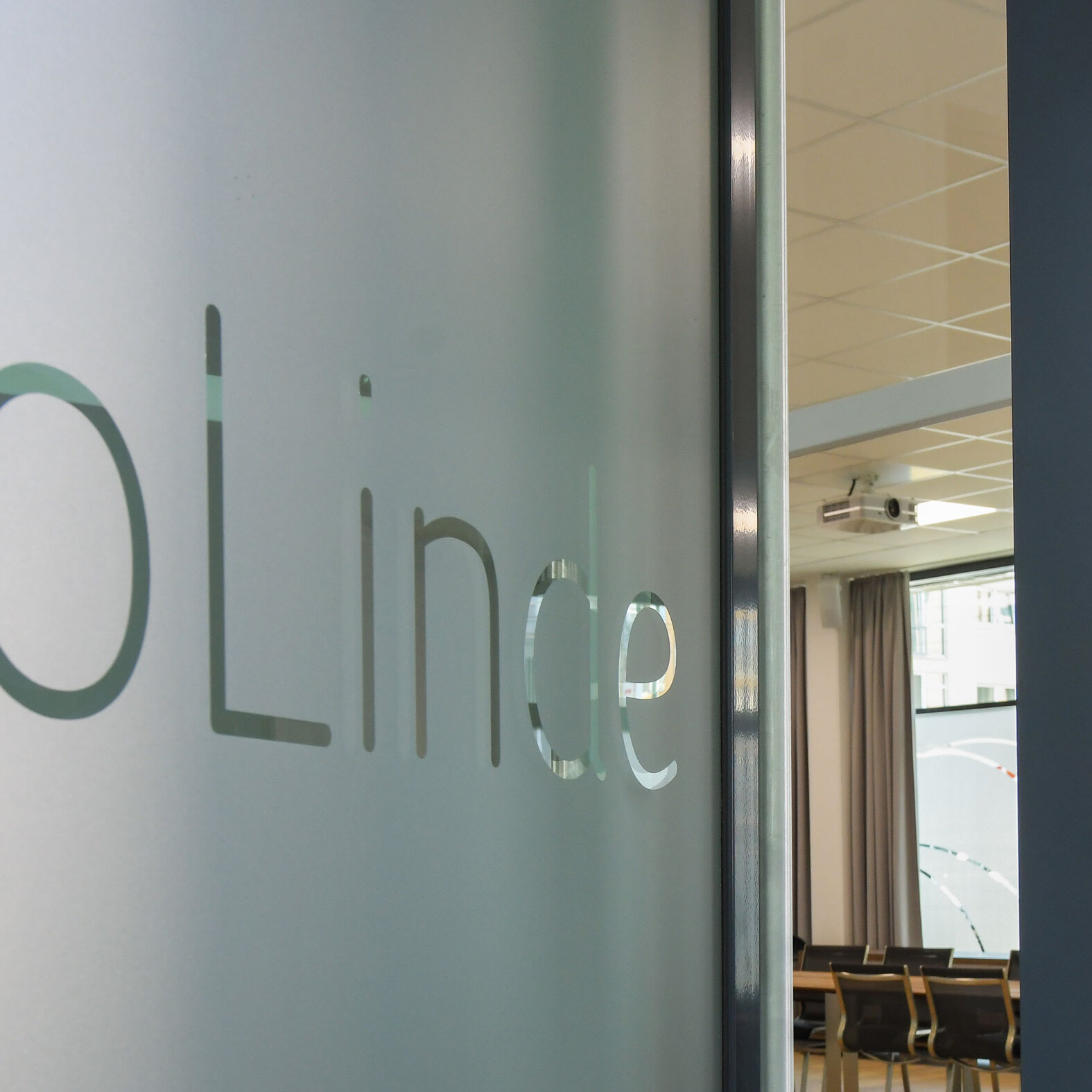 Glaswand mit der Beschriftung „JoLinde“. Durch die geöffnete Tür ist ein Gemeinschaftsraum mit Tischen, Stühlen und Beamer an der Decke zu sehen.