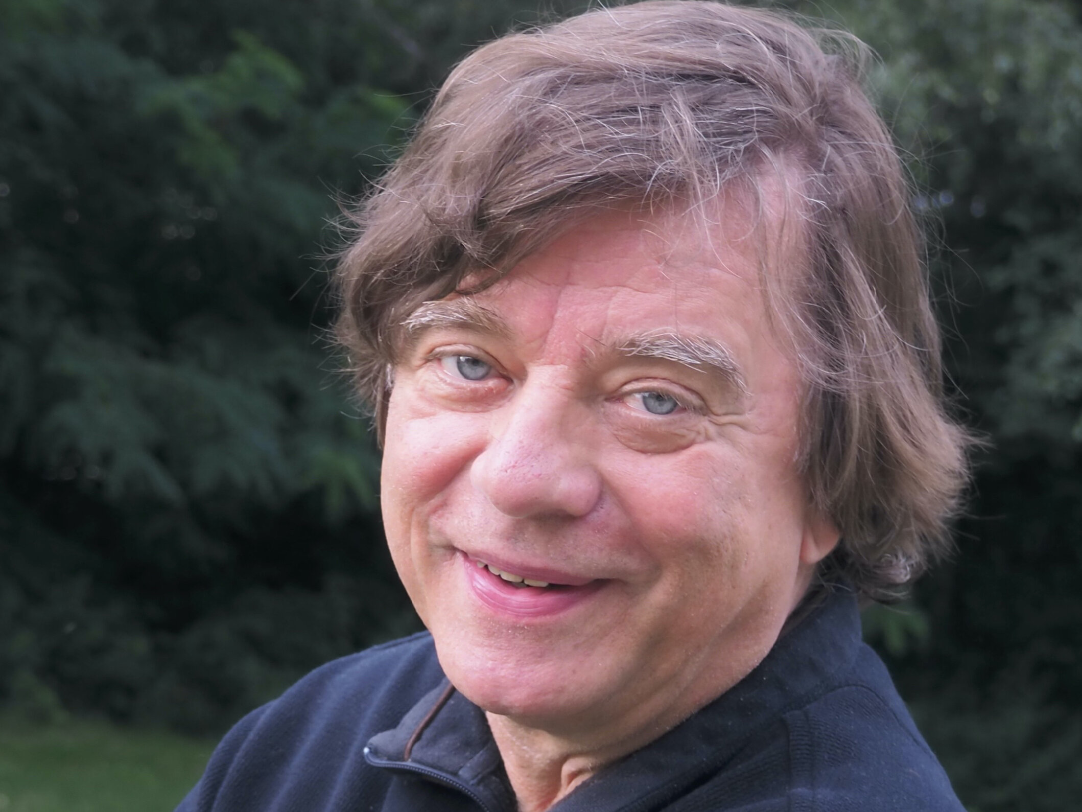 Mann im schwarzen Pullover steht draußen vor einer grünen Hecke und lacht in die Kamera. Er hat die Arme überkreuzt.